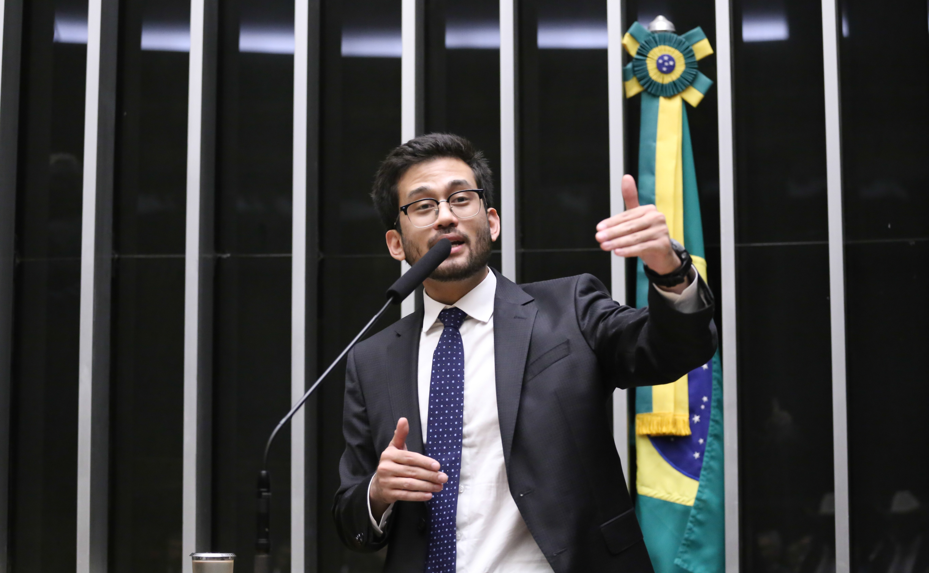 Deputado Kim Kataguiri fala ao microfone. Ele é um homem claro de cabelo escuro. Ele usa um terno preto, camisa branca, gravata azul escuro e óculos. Ao fundo, no lado direito, há uma bandeira do Brasil