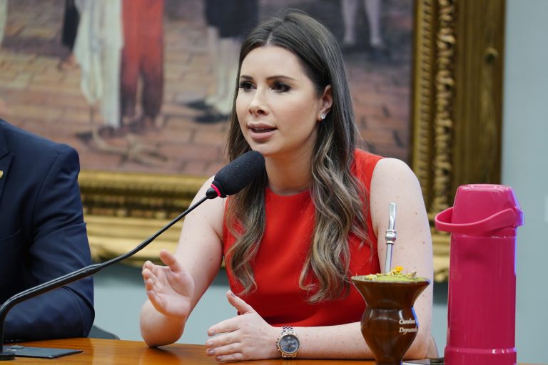 Deputada Caroline de Toni está sentada à mesa onde fala ao microfone