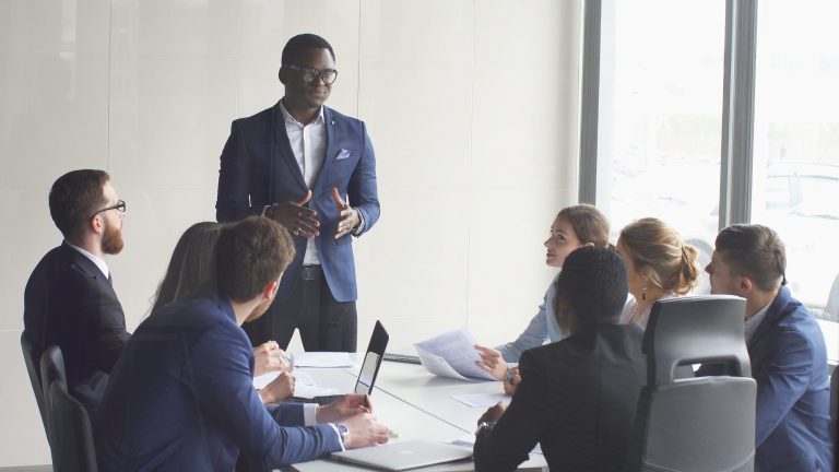 Um homem negro de terno está em pé falando para vários funcionários que estão sentados ao redor de uma mesa