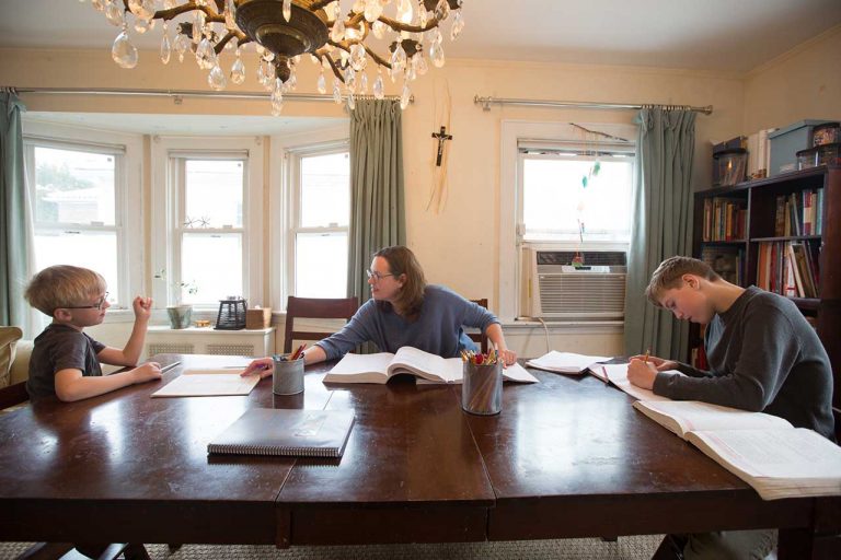 Dois meninos estão sentados à mesa estudando. A mãe está sentada com eles os ensinando
