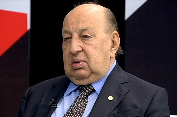 Deputado Simão Sessim está sentado de terno, ao fundo há um painel preto, vermelho e branco