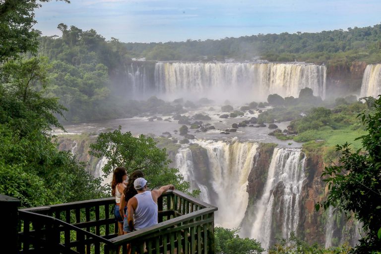 Turistas estão numa ponte olhando uma cachoeira