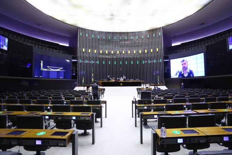 Plenário da Câmara quase vazio com um telão ao fundo por meio do qual os deputados participam remotamente das votações