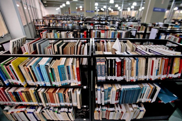 Fotografia em plano médio de uma biblioteca ampla e organizada, com foco em duas estantes pretas de metal repletas de livros coloridos em primeiro plano. Ao fundo, diversas outras fileiras de estantes sob iluminação clara de teto.