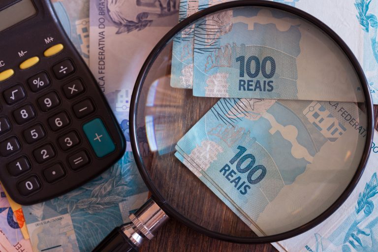 Em cima da mesa há várias notas de dinheiro, uma calculadora e uma lupa