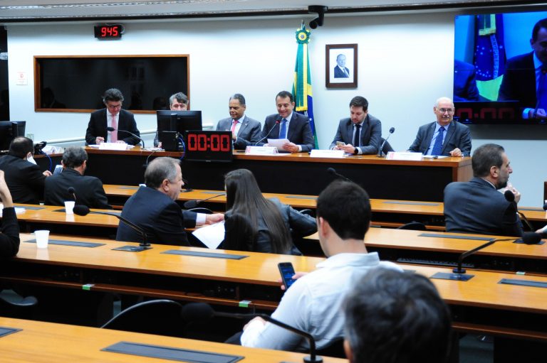 Várias pessoas estão sentadas num plenário ouvindo uma pessoa que fala numa mesa ao fundo