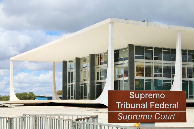Foto de um prédio baixo branco com fachada de vidro num dia nublado. No primeiro plano aparece uma placa escrito: Supremo Tribunal Federal
