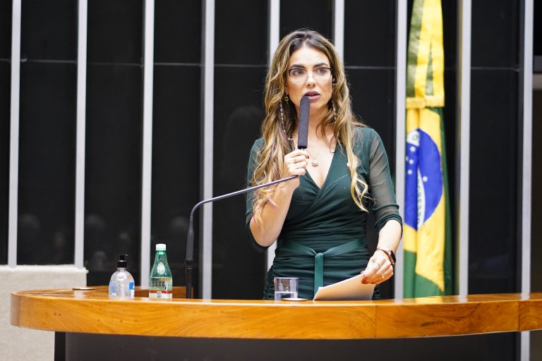 Deputada Paula Belmonte fala ao microfone. Ela é branca, tem o cabelo comprido e loiro e usa um vestido escuro