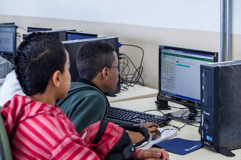 Fotografia de dois jovens negros sentados em um laboratório de informática, vistos de perfil. Eles olham atentamente para o monitor de um computador que exibe uma interface de edição de texto ou código.