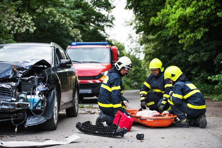 Uma mulher está deitada numa maca numa estrada. Os bombeiros estão ajoelhados ao lado dela. Ao lado um carro batido na frente