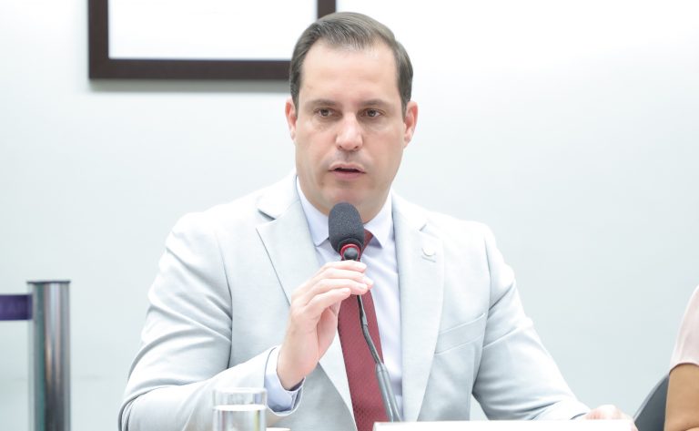 Deputado Nicoletti fala ao microfone. Ele é claro, tem o cabelo castanho, veste um terno bege com gravata avermelhada