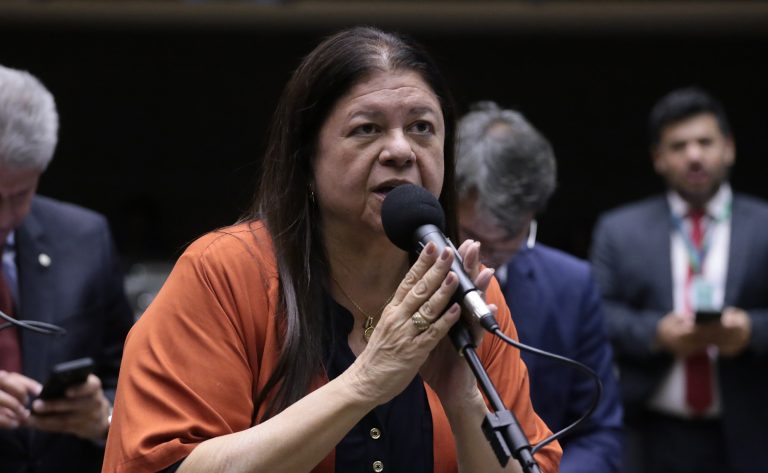 Deputada Laura Carneiro fala ao microfone. Ela é branca, tem o cabelo comprido e escuro, veste uma blusa laranja